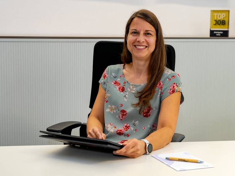 Julia Hildebrand sitzt lächelnd  in einem geblümten Kleid an einem Tisch mit einem Tablet in der Hand und einem Notizblock sowie Stift vor sich.