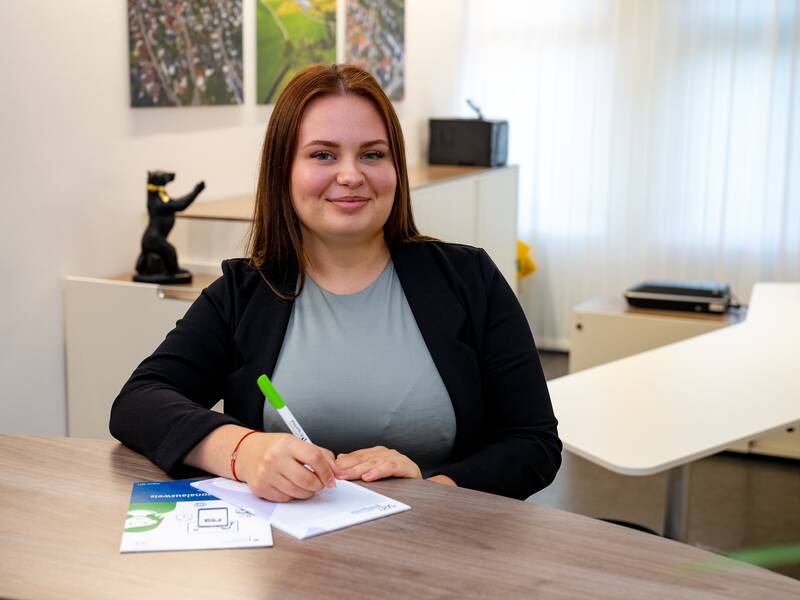 Lilia Hrusc sitzt lächelnd an einem Schreibtisch und schreibt mit einem Stift auf ein Blatt Papier.