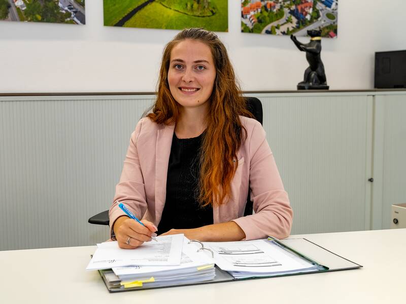 Anna Knop in einem rosa Blazer sitzt an einem Schreibtisch mit einem geöffneten Ordner und einigen Papieren, während sie mit einem Stift schreibt und lächelt.