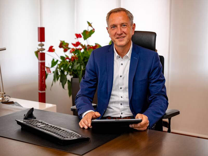 Bürgermeister Michael Schmitt in einem blauen Anzug sitzt lächelnd an einem Schreibtisch und hält ein Tablet, während im Hintergrund Pflanzen zu sehen sind.