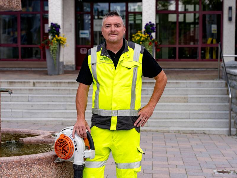 Matthias Kleiser in gelber Arbeitskleidung steht mit einer Laubbläsermaschine neben einem Brunnen vor einem Gebäude mit Treppen und Blumenschmuck.