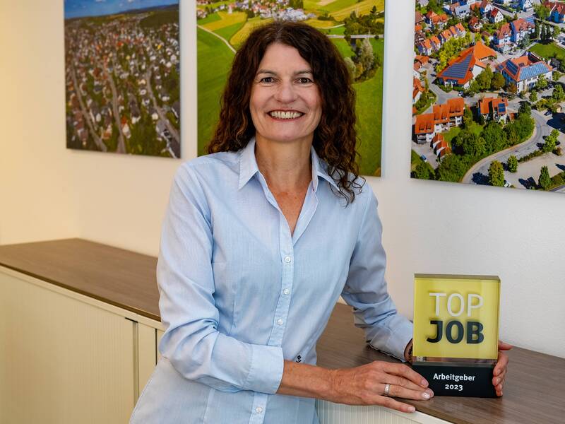 Elke Dufner in einem hellblauen Hemd lächelt und lehnt sich an ein Sideboard, während sie eine Auszeichnung mit der Aufschrift "Top Job Arbeitgeber 2023" hält, im Hintergrund sind Luftaufnahmen von Landschaften und Gebäuden zu sehen.