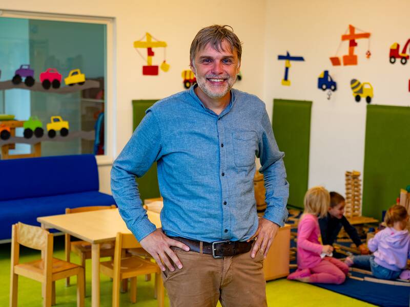 Rainer Kruspel in blauem Hemd steht lächelnd in einem bunten Kindergarten, während Kinder im Hintergrund spielen.