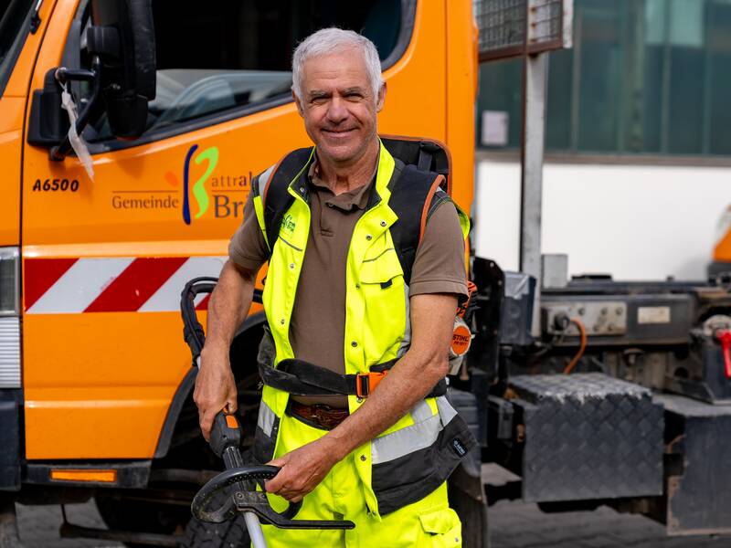 George Pena in gelber Warnkleidung steht lächelnd vor einem orangefarbenen Kommunalfahrzeug.