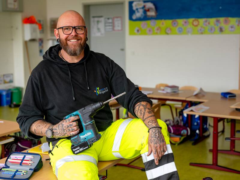 Ralf Dutty  mit Bart und Brille sitzt lächelnd in einem Klassenzimmer auf einem Tisch und hält eine Bohrmaschine in der Hand; er trägt eine schwarze Kapuzenjacke und gelbe Arbeitshosen.