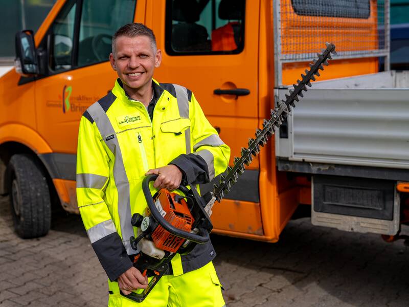 Sascha Häfle in gelber Warnschutzkleidung hält eine Heckenschere und steht vor einem orangefarbenen Fahrzeug.
