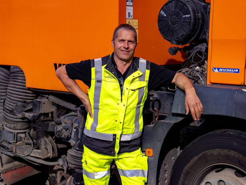Gerhard Schleicher in leuchtend gelber Arbeitskleidung steht lächelnd neben einem großen orangefarbenen Fahrzeug.