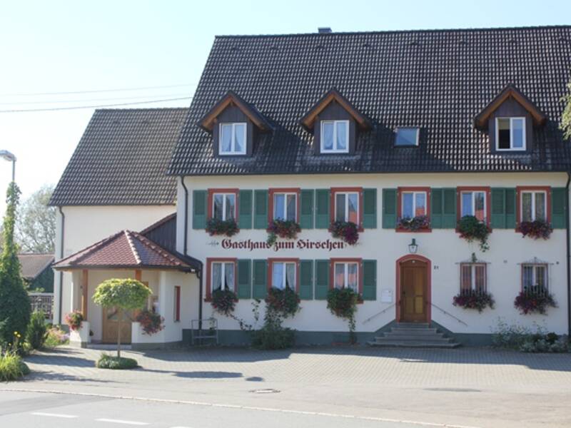 Das Bild zeigt das traditionelle Gasthaus  Kirschen mit Holzläden an den Fenstern und Blumenkästen, das neben einer Straße liegt.
