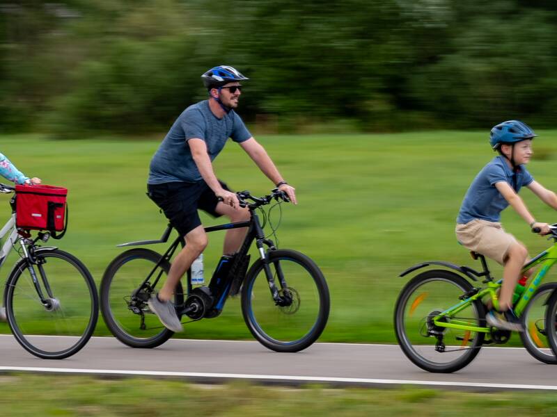 Eine Familie fährt gemeinsam mit Fahrrädern und einem Kindersitz auf einem Radweg durch eine grüne Landschaft.