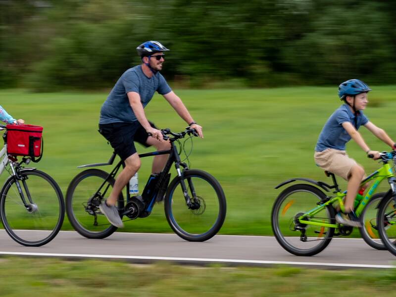 Eine Familie fährt gemeinsam mit Fahrrädern und einem Kindersitz auf einem Radweg durch eine grüne Landschaft.